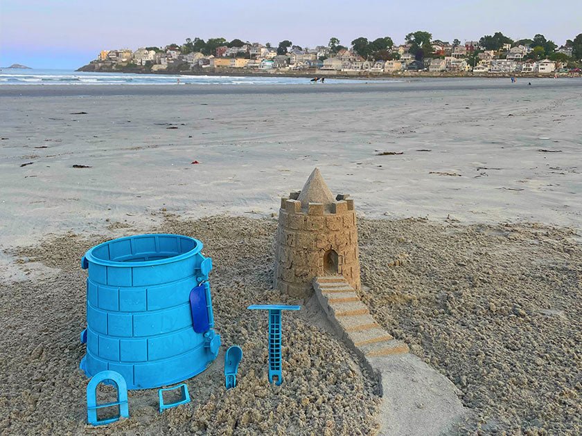 Blue Basic Tower mold and detailing tools beside a pointed sandcastle tower and staircase on the beach.