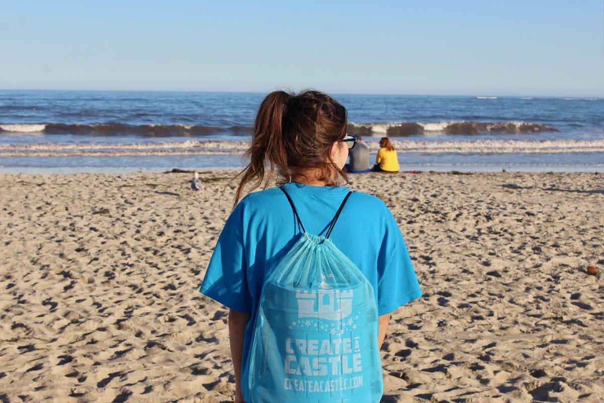 Rear view of a person wearing a blue Create A Castle drawstring backpack on the beach.