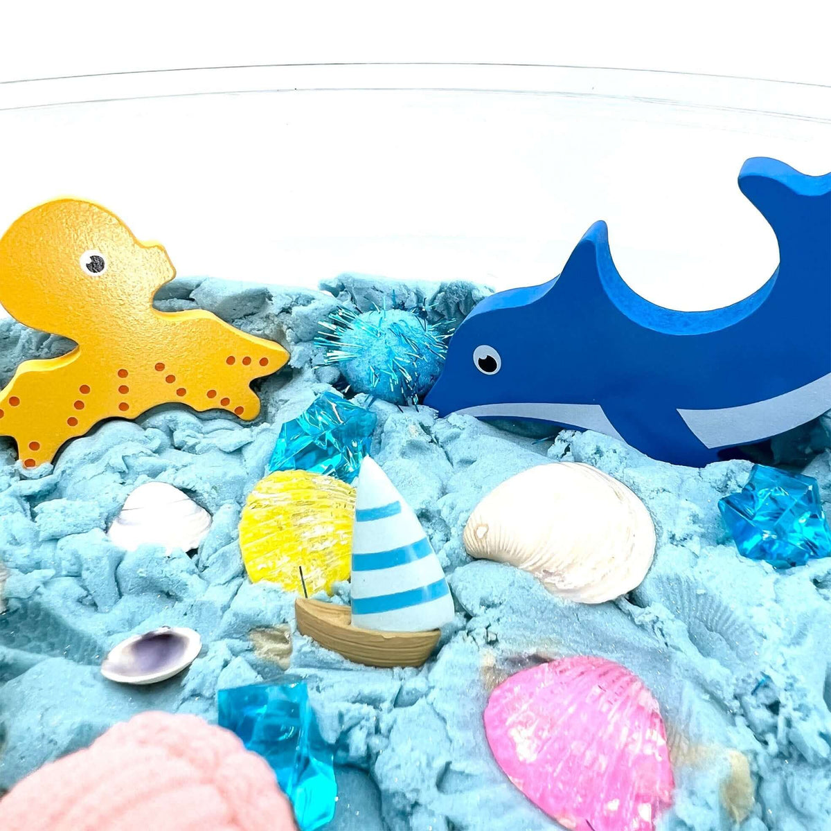 Octopus and dolphin figures beside shells and a miniature sailboat in blue sand