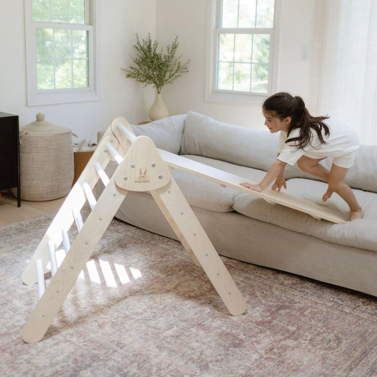 Child crawling on a ramp bridging a white-rung climbing triangle and a sofa.
