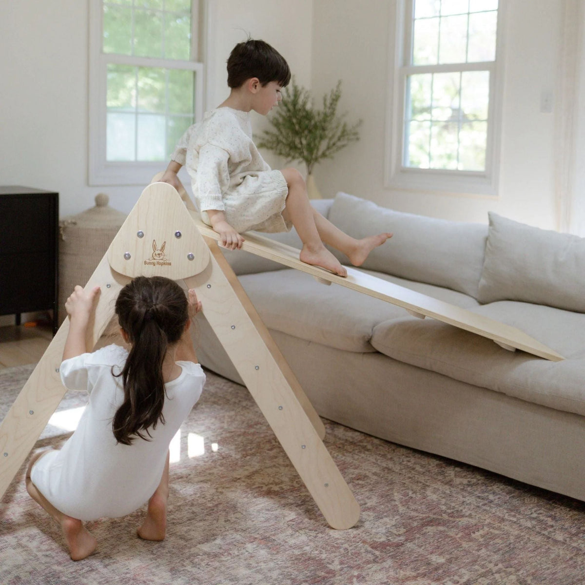 One child sitting on a ramp between a white-rung triangle and sofa while another reaches toward the triangle.
