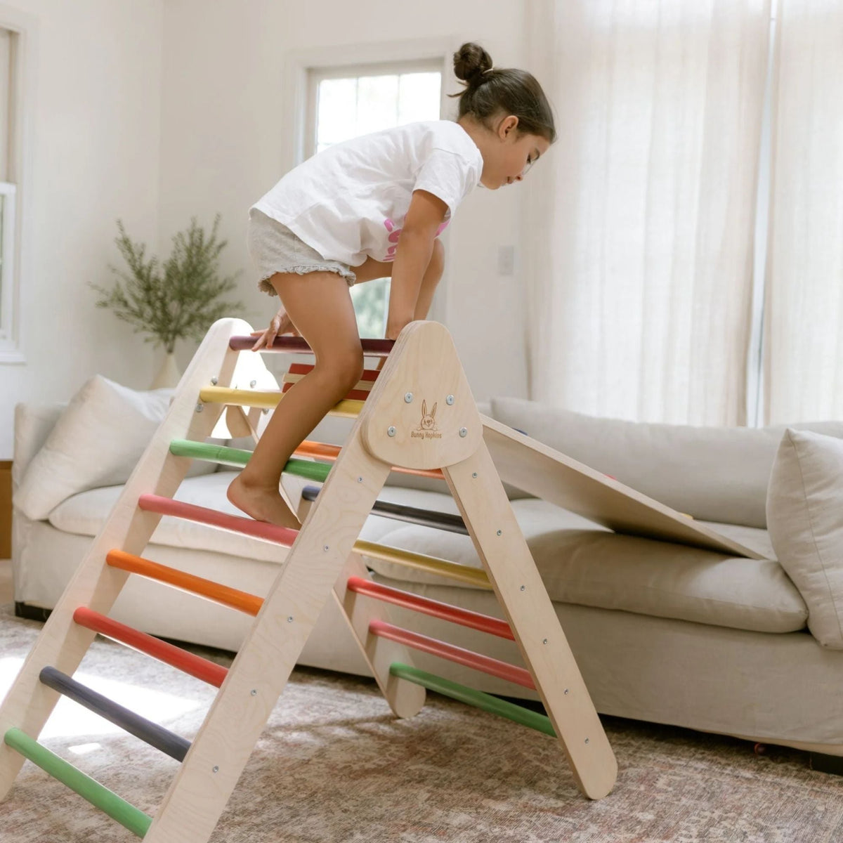Child climbing over a rainbow-rung triangle beside a sofa.