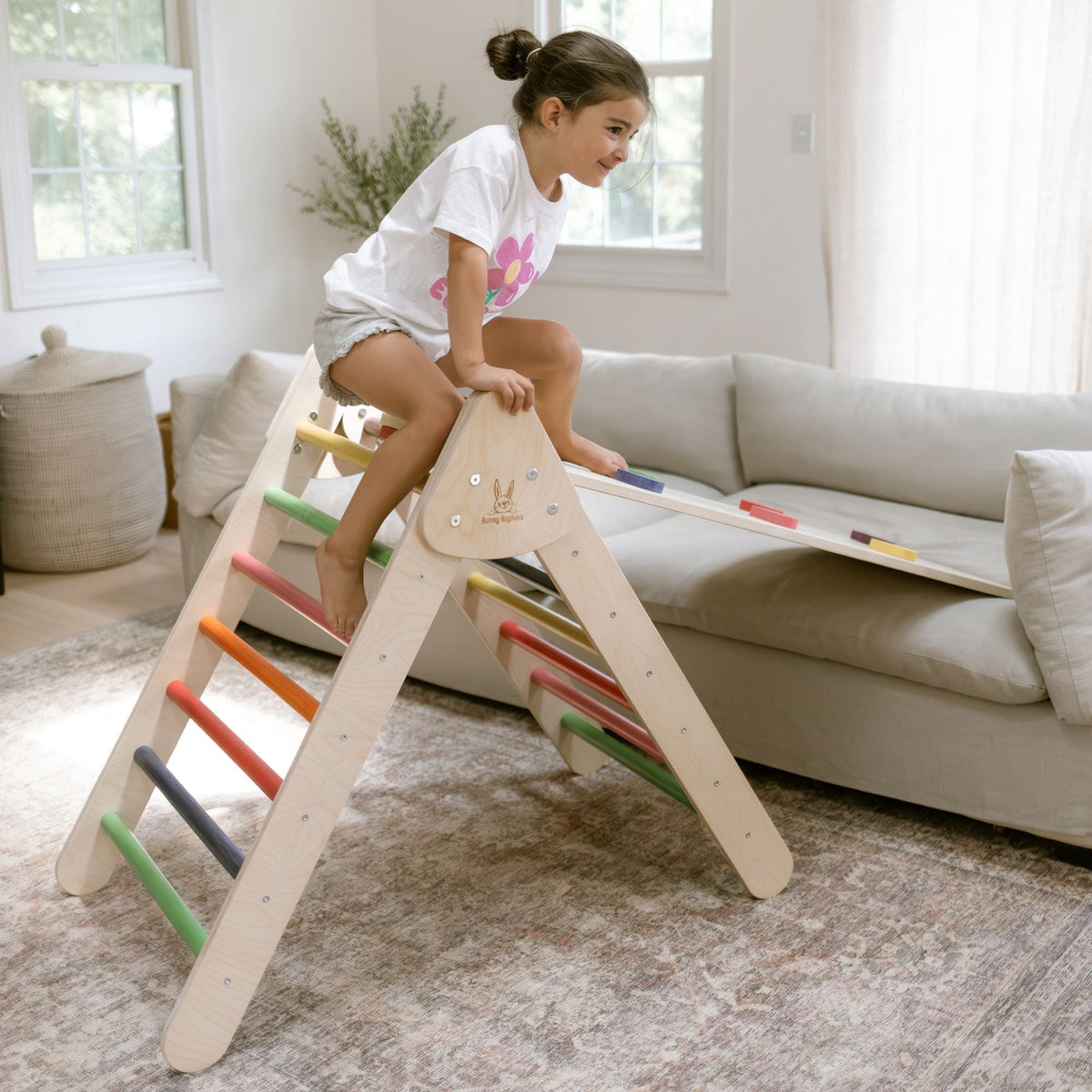 Child sitting across the top of a rainbow-rung climbing triangle, with a ramp leading to a sofa.