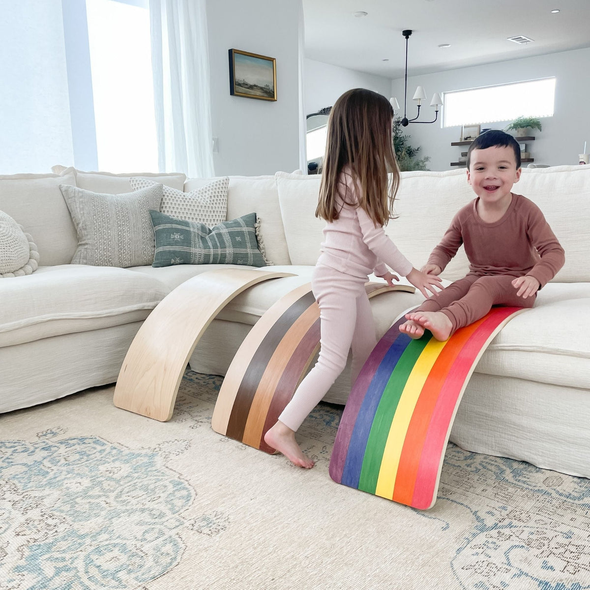 Two children playing with three curved wobble boards propped against a sofa, including a rainbow board.