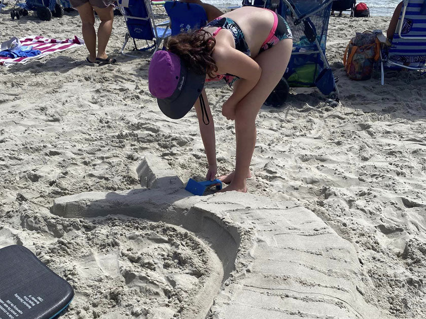 Person using a blue sculpting tool to smooth a large curved sand creation on the beach.