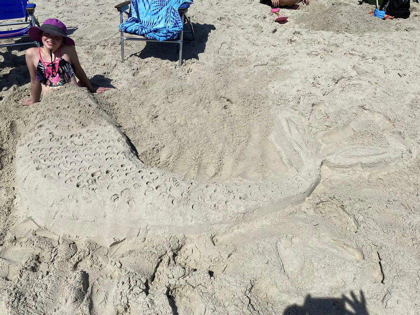 Person sitting at the head of a large mermaid-tail sand sculpture on the beach.