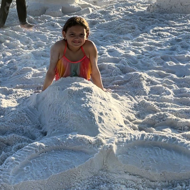Person sitting behind a sand mermaid tail with two broad fins on a sunny beach.