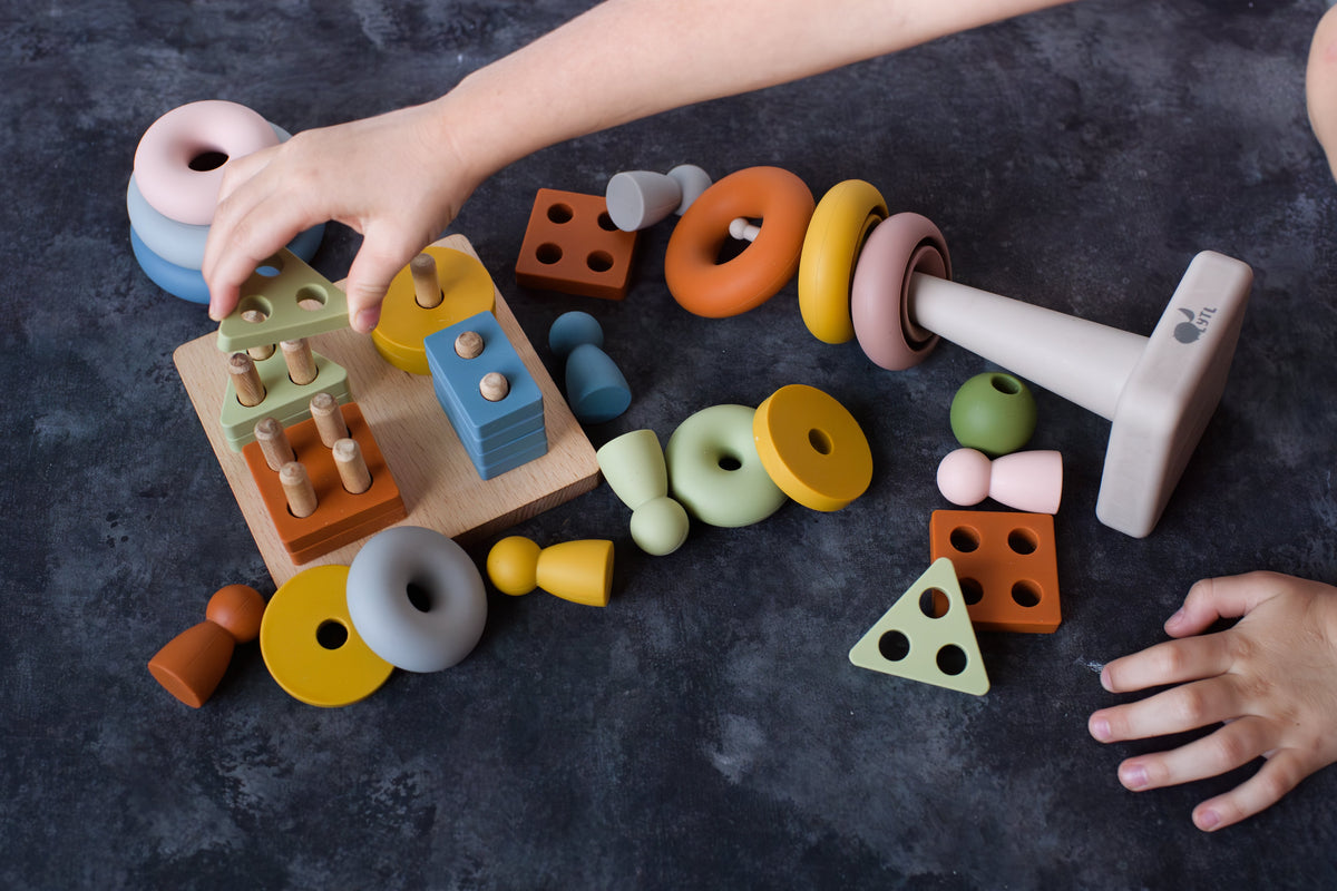 Hands reaching toward peg dolls, shape puzzle pieces, and rings on a dark tabletop.