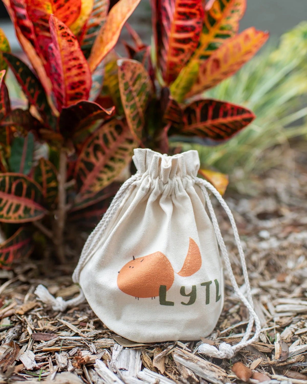 LYTL drawstring toy bag standing on wood chips in front of colorful foliage.