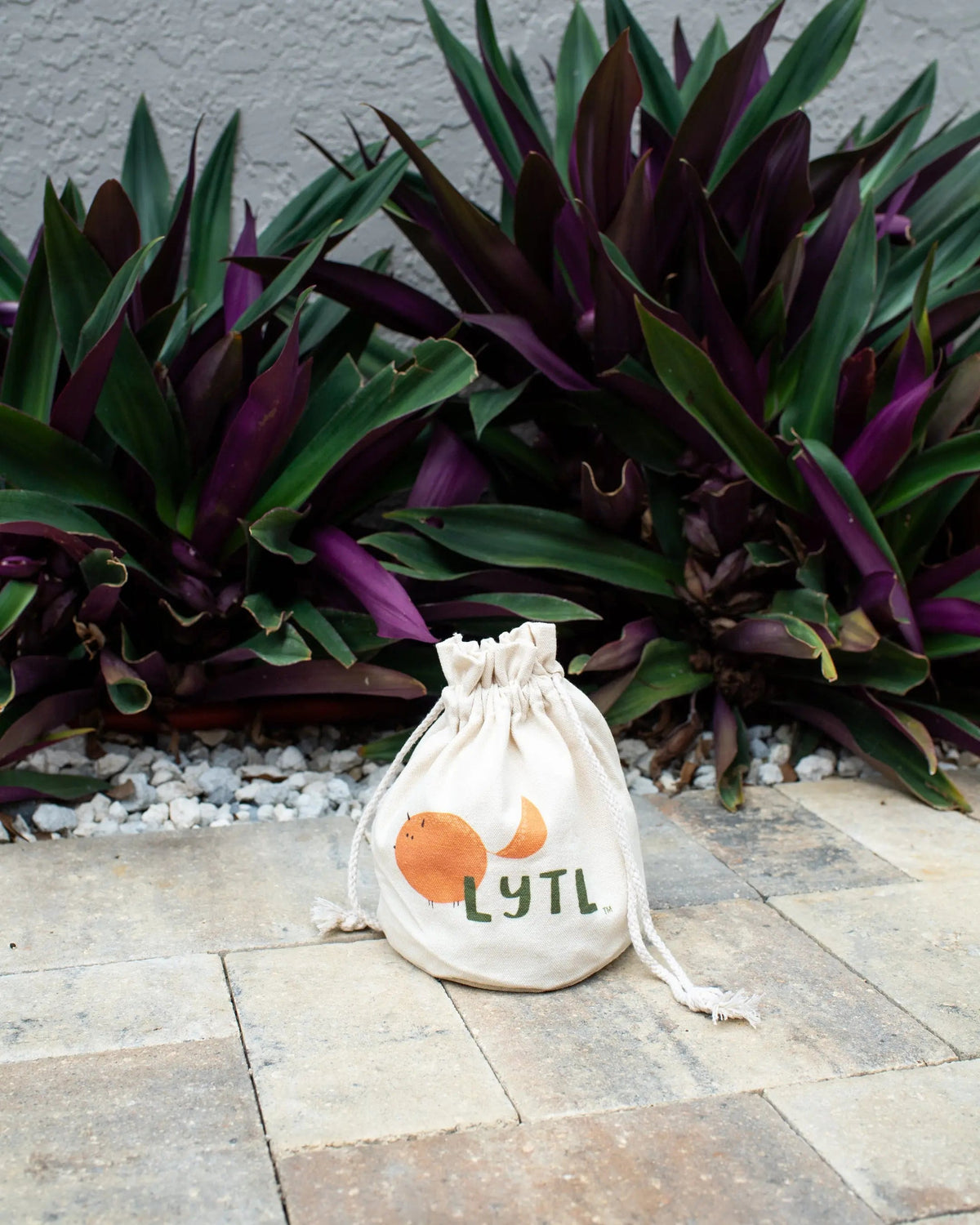 Natural canvas LYTL toy bag on paving stones in front of purple and green plants.