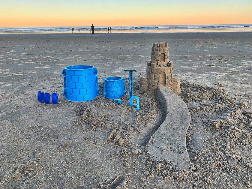 Two blue tower molds, removable buckles and detailing tools beside a tiered sandcastle and staircase at sunset.