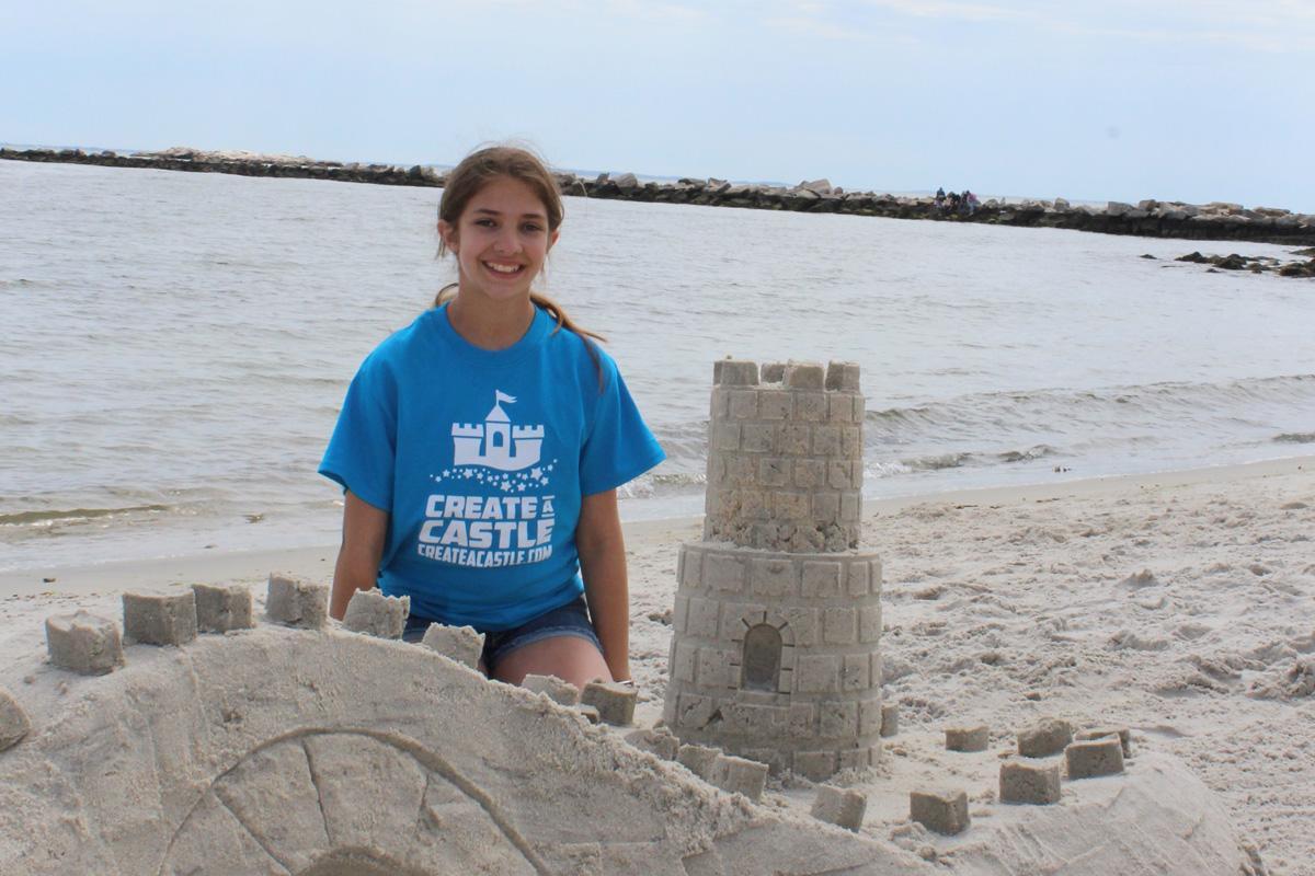 Person in a blue Create A Castle shirt behind a sand bridge and two-tier sandcastle at the shoreline.