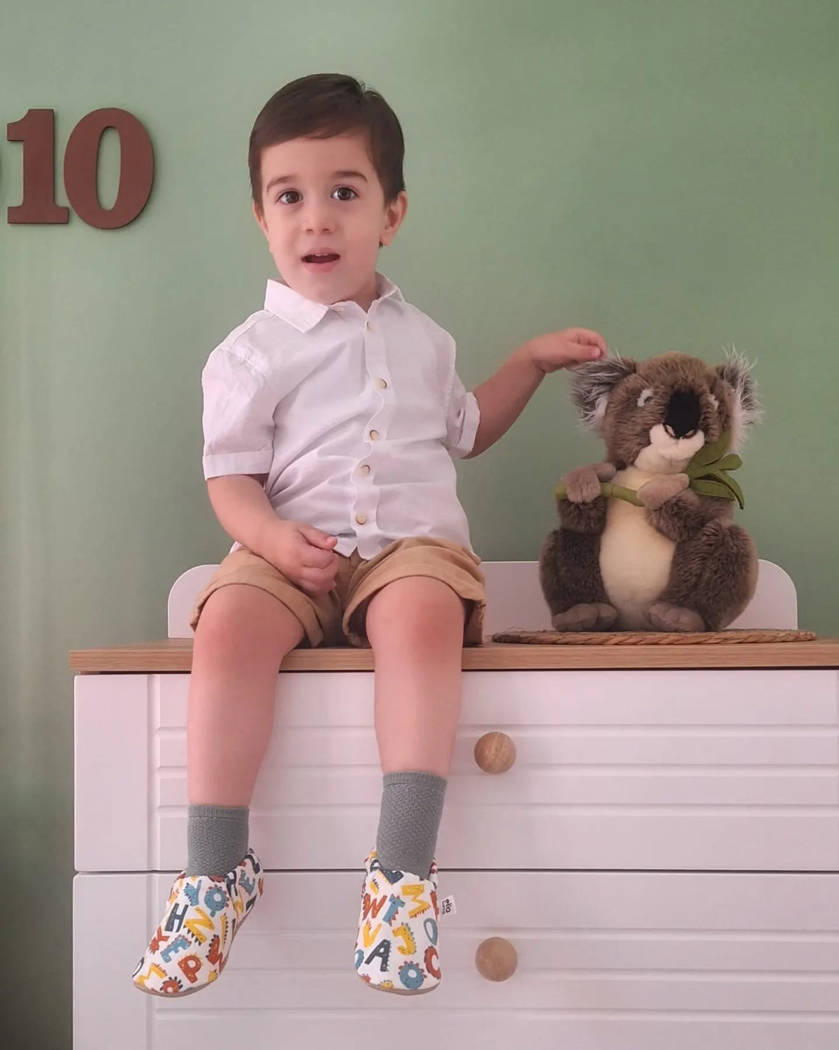 Child seated on a dresser wearing alphabet-print moccasins, front view.