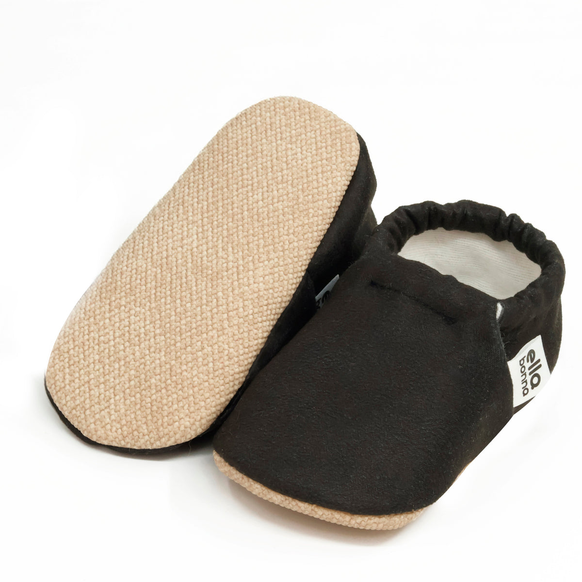 Black slip-on baby moccasins with cream lining and tan soles, with one sole facing the camera on a white background.