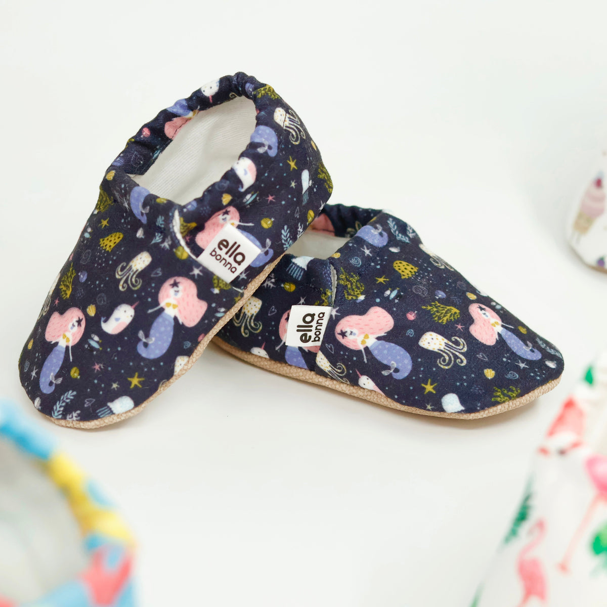 Side views of navy slip-on baby moccasins with mermaids and sea-creature print on a white background.