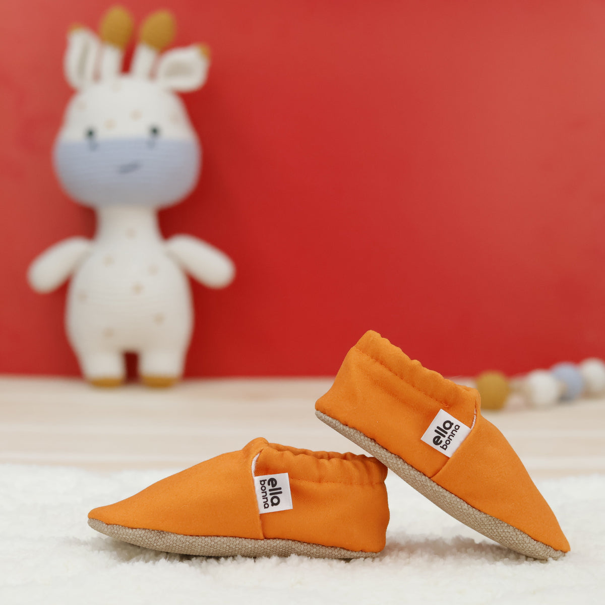 Orange slip-on baby moccasins with cream lining and tan soles arranged together to show the sides and uppers.
