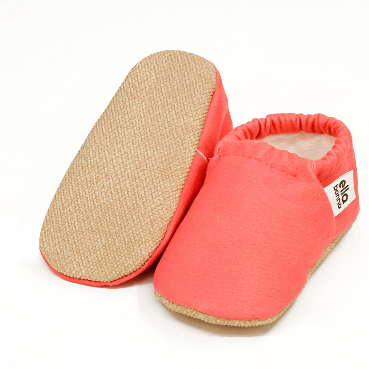 Coral pink slip-on baby moccasins with cream lining and tan soles, with one sole facing the camera on a white background.