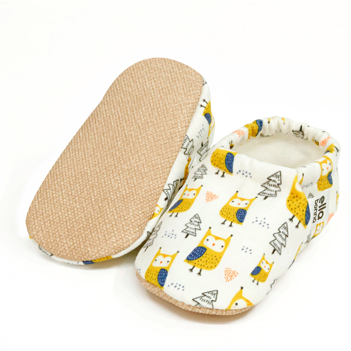 Cream slip-on baby moccasins with yellow owl and tree print, with one sole facing the camera on a white background.