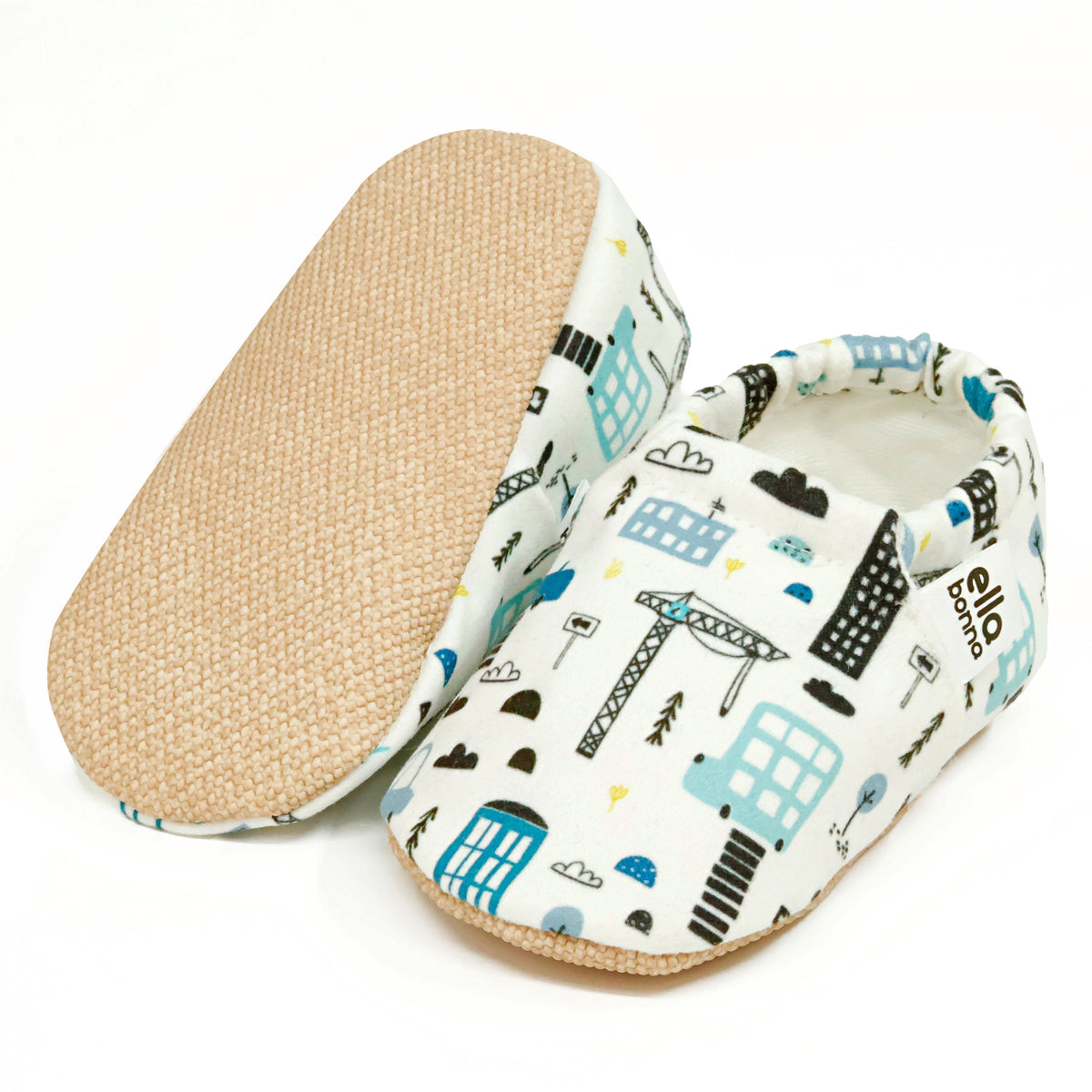 Cream slip-on baby moccasins with blue-and-black city building print, with one sole facing the camera on a white background.