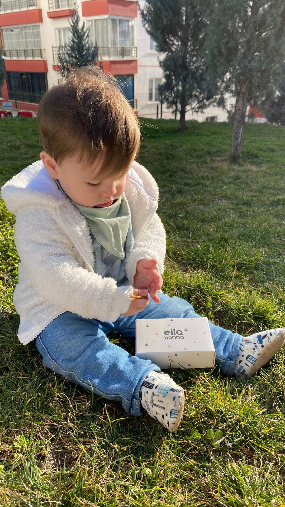Child seated on grass holding an Ella Bonna card and wearing city-print moccasins.