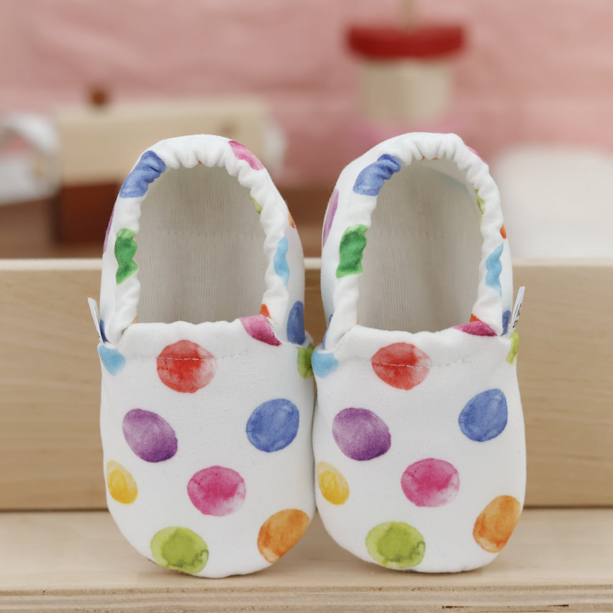 Pair of white slip-on baby moccasins with multicolored watercolor dot print, showing the openings and uppers.
