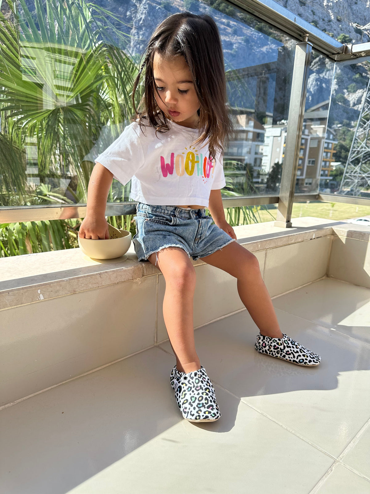 Child seated on a window ledge wearing leopard-print moccasins.