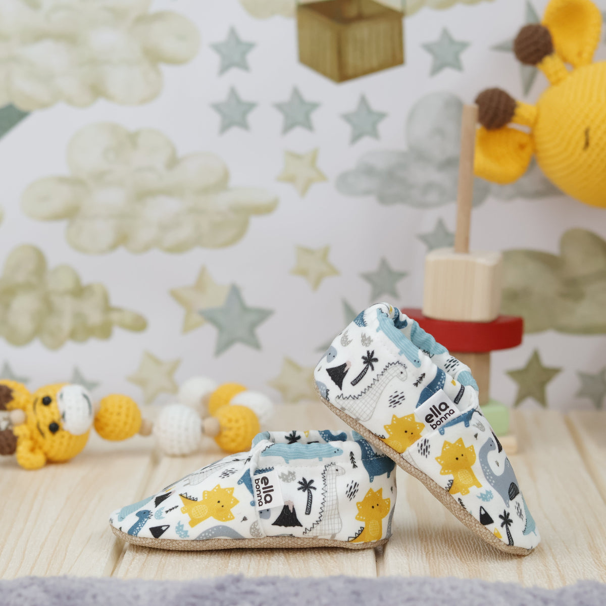 Cream slip-on baby moccasins with blue, gray and yellow dinosaur print arranged together to show the sides and uppers.