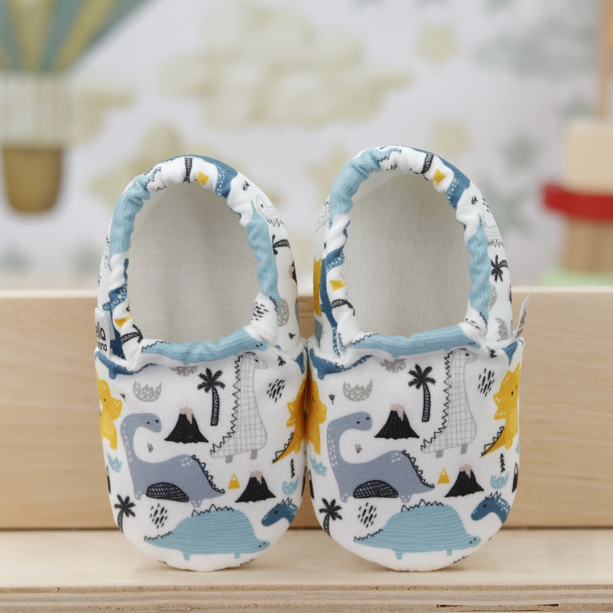 Pair of cream slip-on baby moccasins with blue, gray and yellow dinosaur print, showing the openings and uppers.