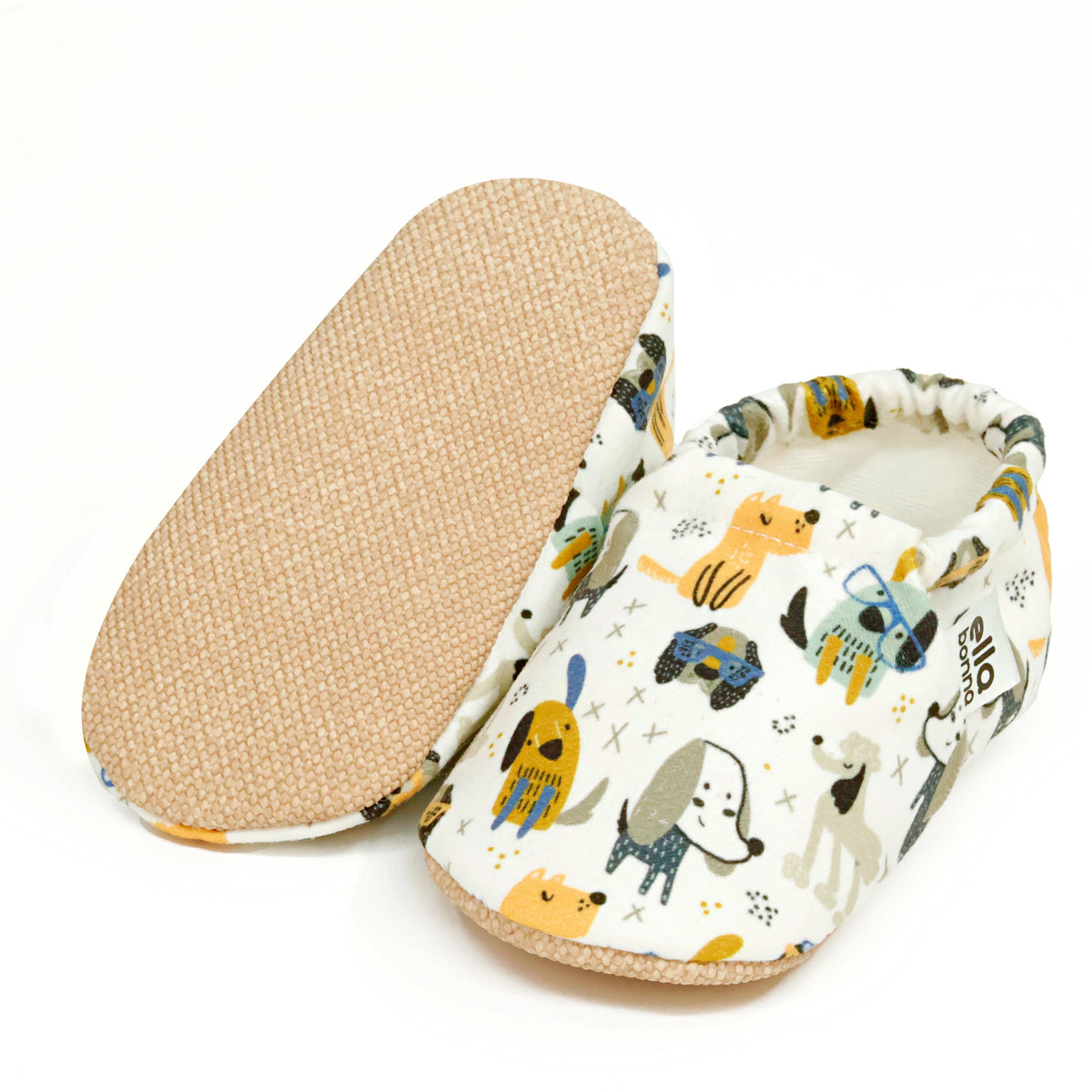 Cream slip-on baby moccasins with yellow, blue and gray dog print, with one sole facing the camera on a white background.
