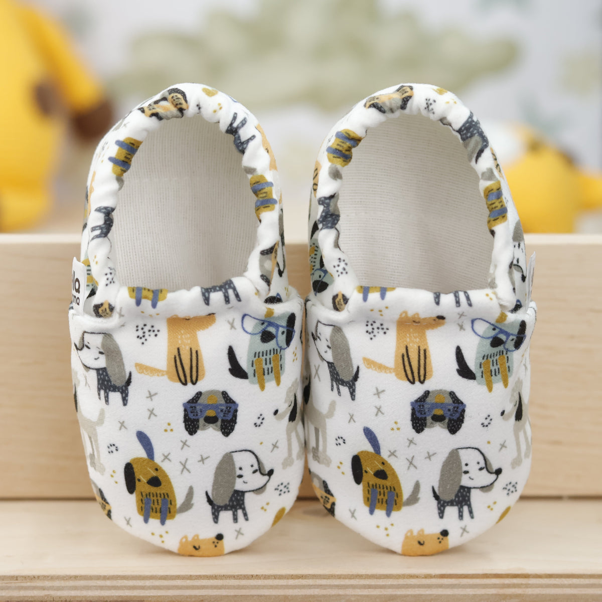 Pair of cream slip-on baby moccasins with yellow, blue and gray dog print, showing the openings and uppers.