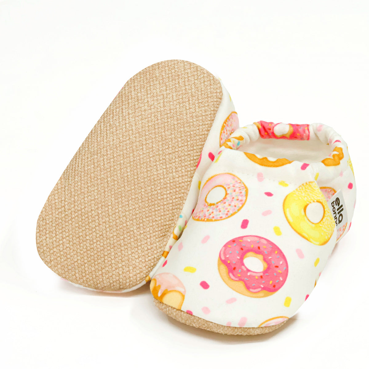 Cream slip-on baby moccasins with pink-and-yellow doughnut print, with one sole facing the camera on a white background.