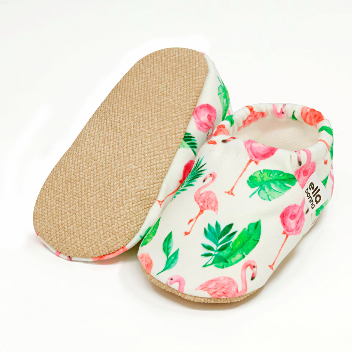 Cream slip-on baby moccasins with pink flamingos and green leaves, with one sole facing the camera on a white background.