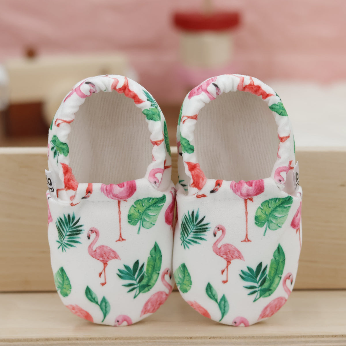 Pair of cream slip-on baby moccasins with pink flamingos and green leaves, showing the openings and uppers.
