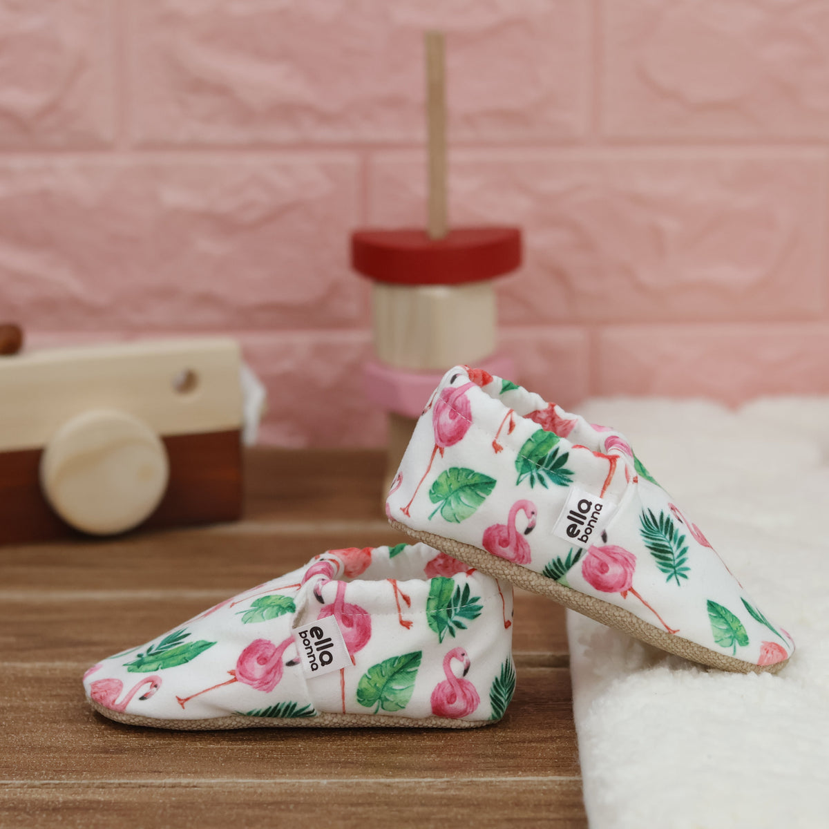 Cream slip-on baby moccasins with pink flamingos and green leaves arranged together to show the sides and uppers.