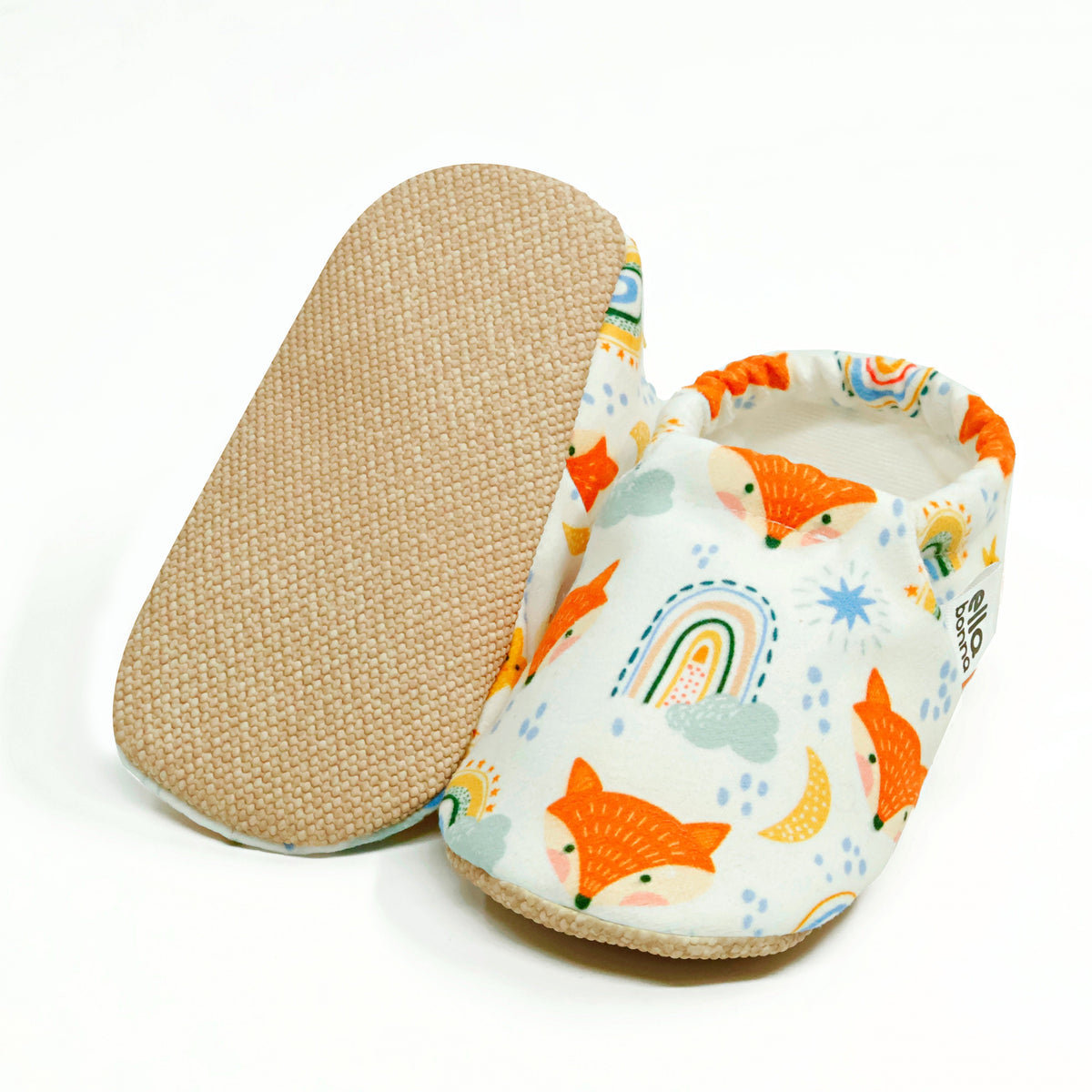 Cream slip-on baby moccasins with orange foxes and rainbows, with one sole facing the camera on a white background.