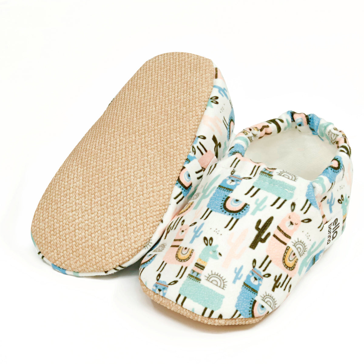 Cream slip-on baby moccasins with blue-and-tan llama and cactus print, with one sole facing the camera on a white background.