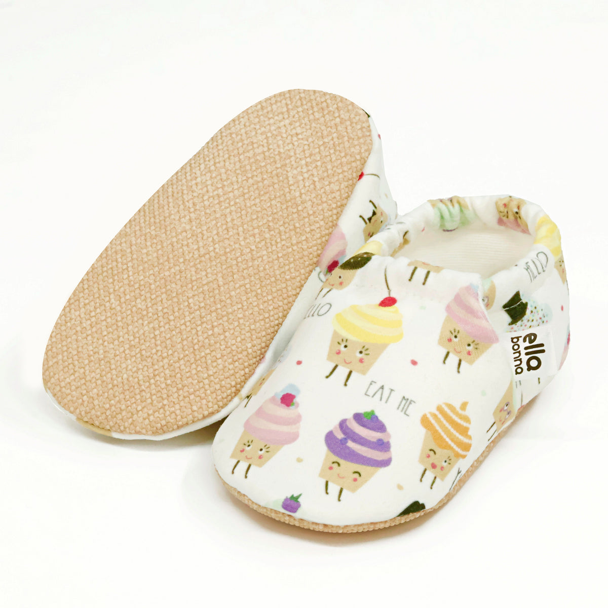 Cream slip-on baby moccasins with pastel cupcake print, with one sole facing the camera on a white background.