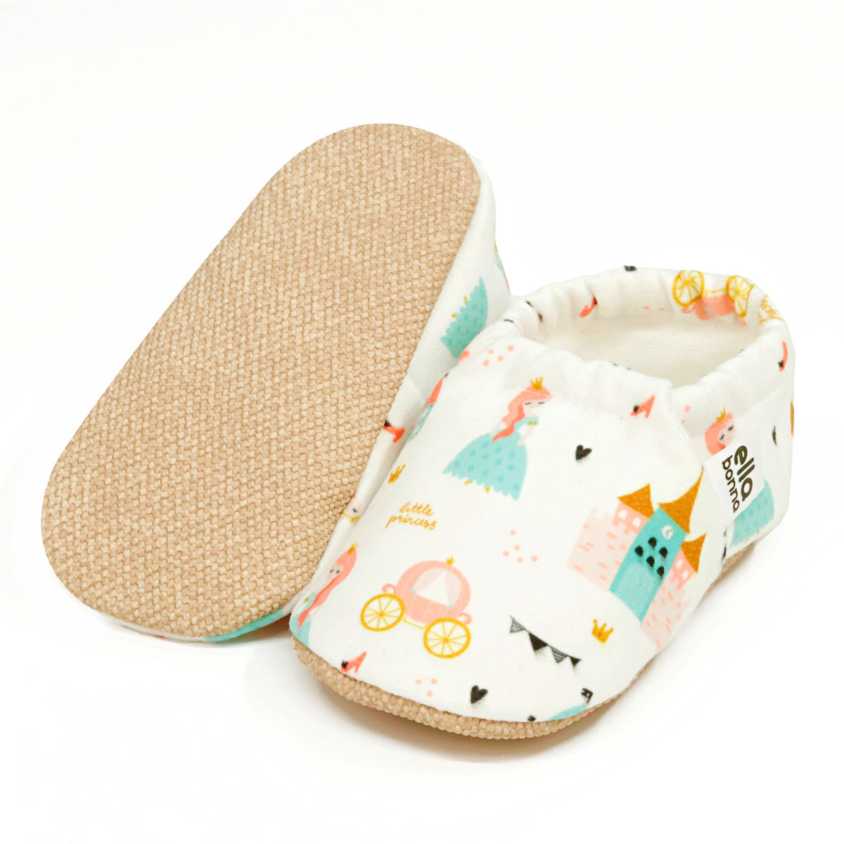 Cream slip-on baby moccasins with princess, castle and carriage print, with one sole facing the camera on a white background.