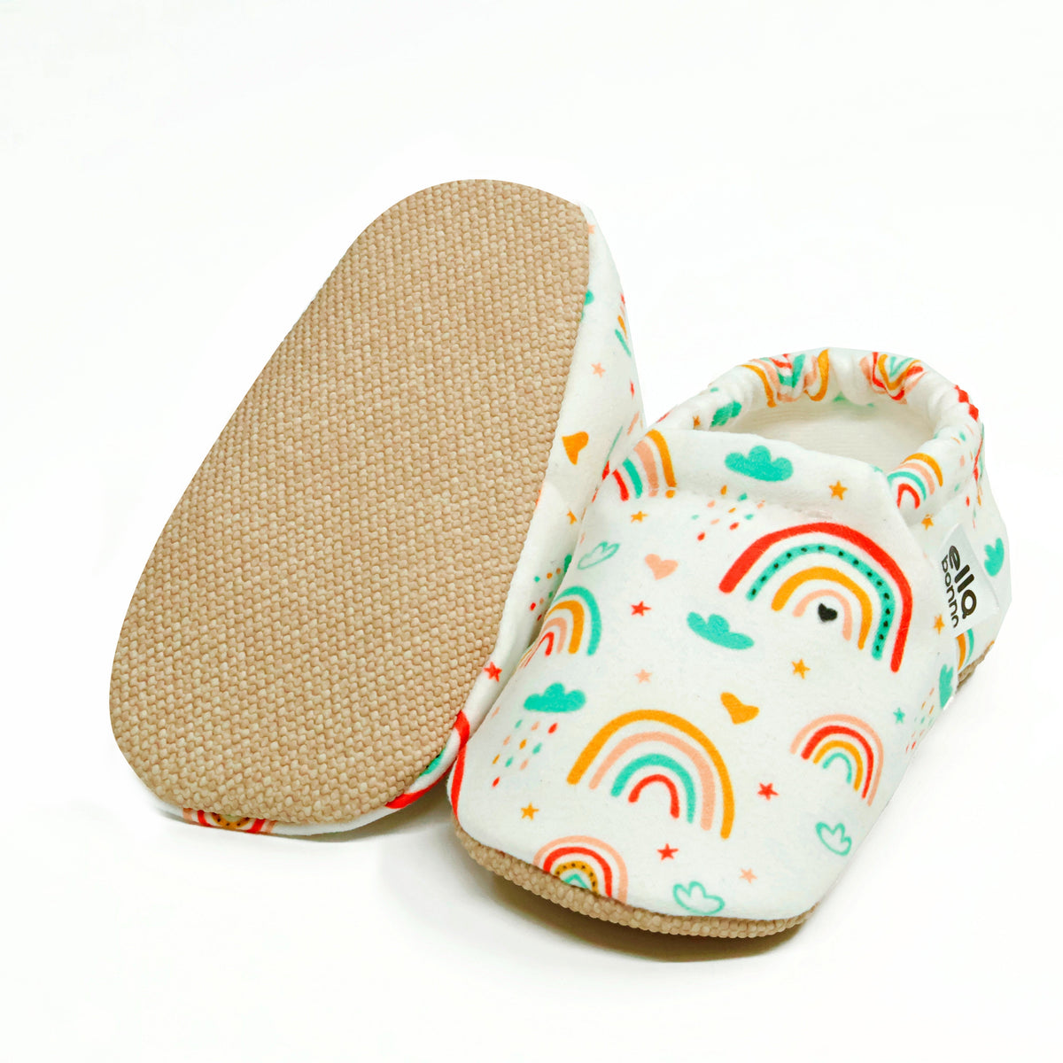 Cream slip-on baby moccasins with colorful rainbow print, with one sole facing the camera on a white background.
