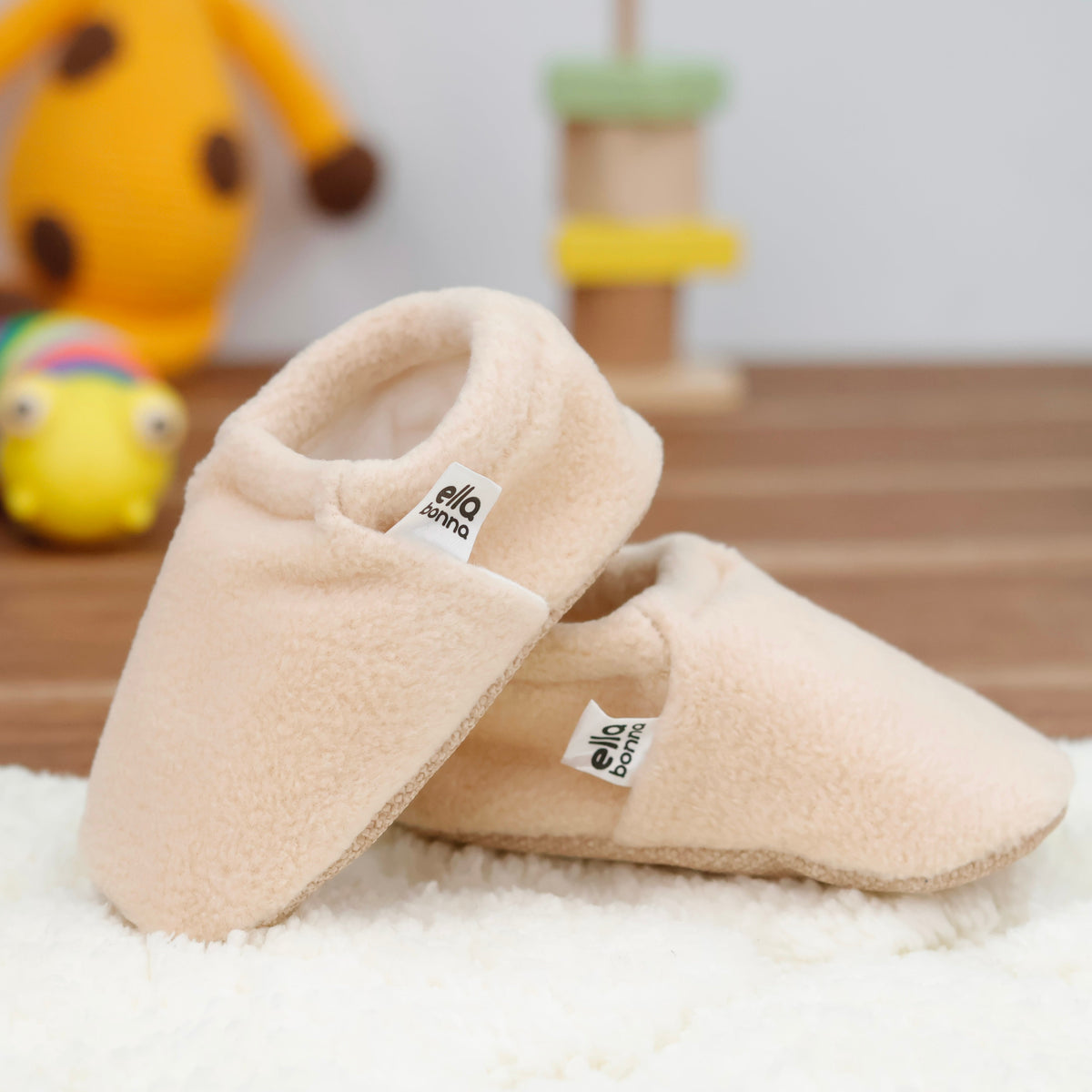 Cream fleece slip-on baby moccasins with tan soles arranged on a surface with one sole facing the camera.