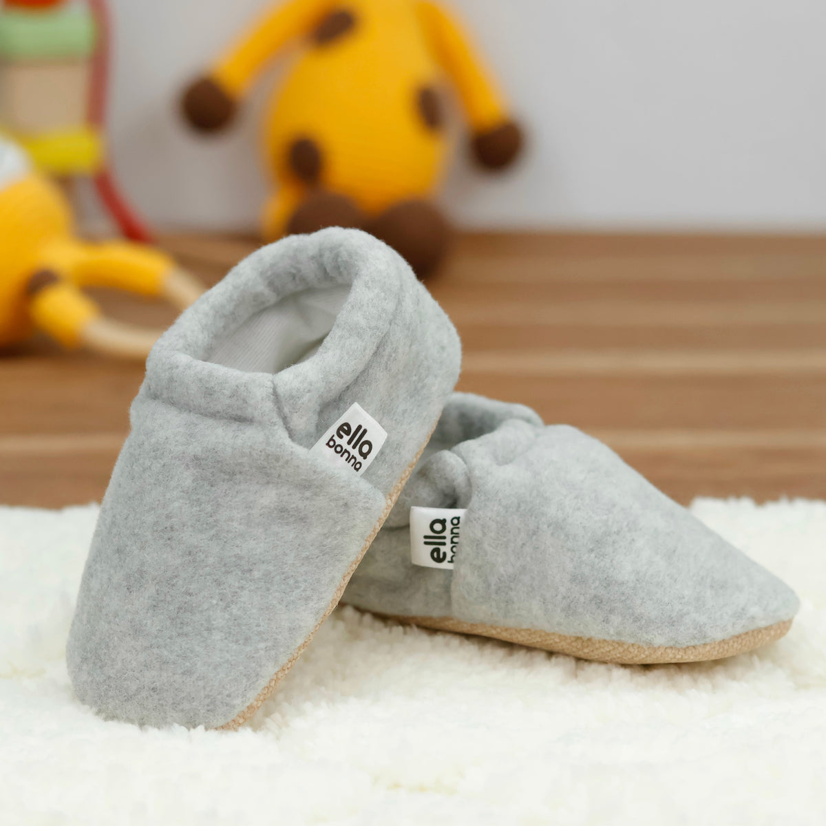 Light gray fleece slip-on baby moccasins with tan soles arranged on a surface with one sole facing the camera.