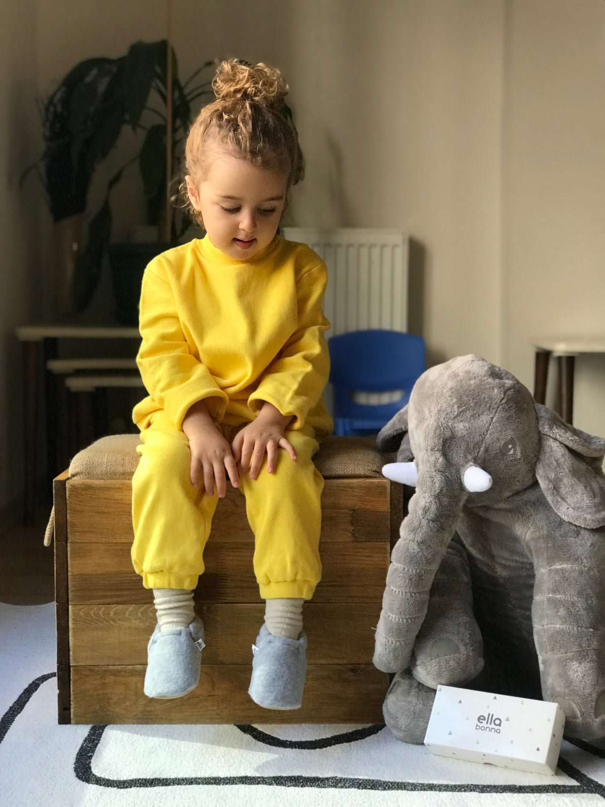 Child seated beside an elephant toy wearing gray fleece moccasins.