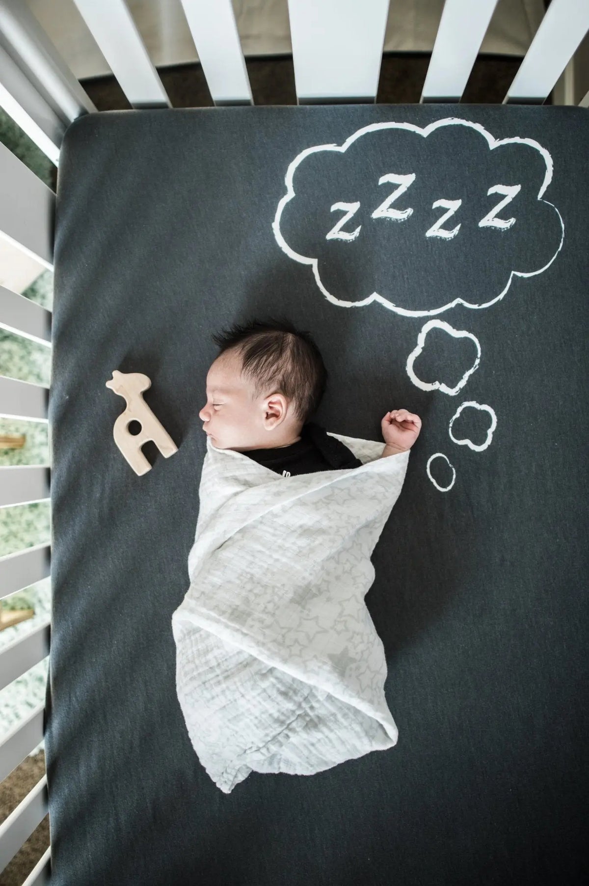 Staged nursery photo with a swaddled baby and a wooden giraffe grasping toy