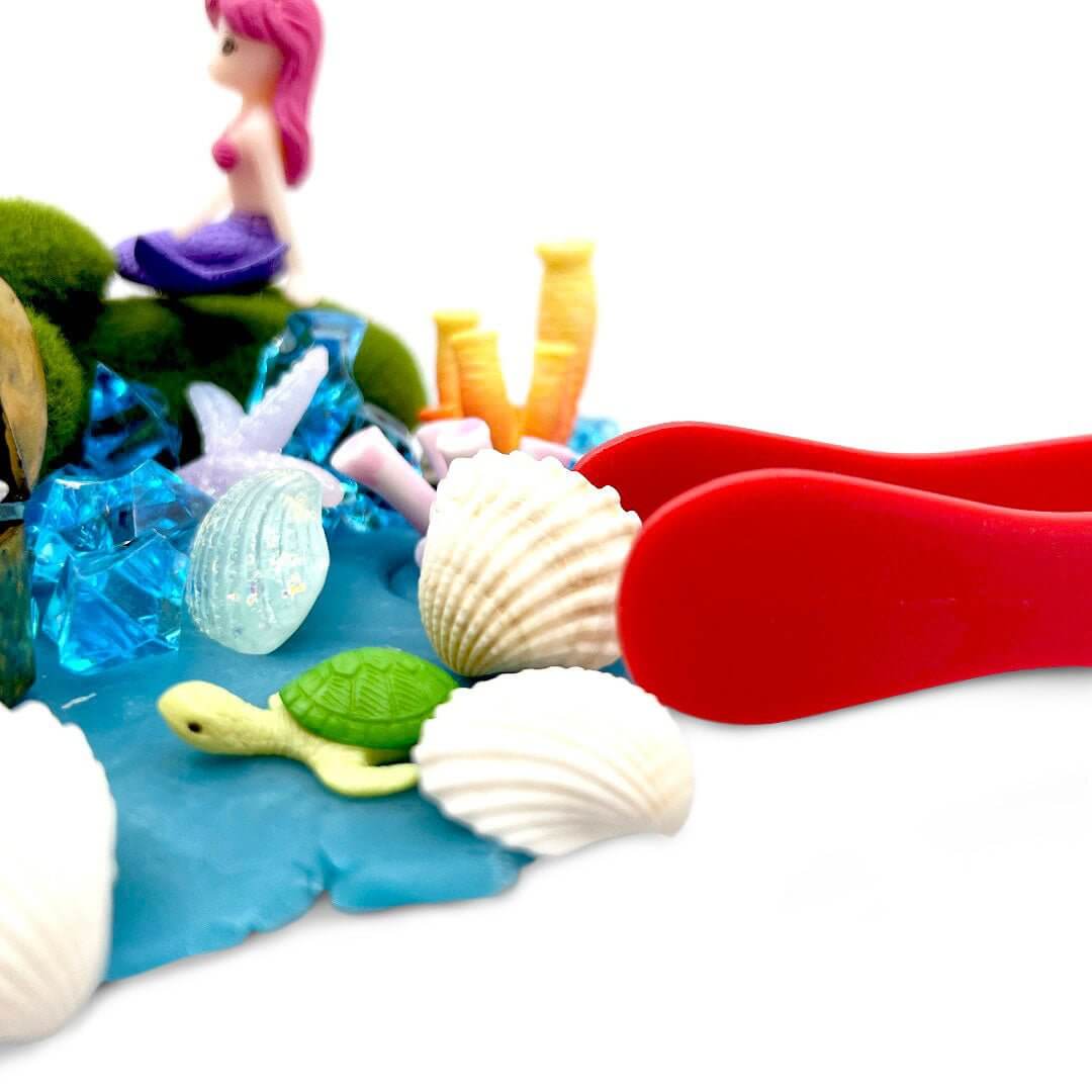 Red tongs beside shells and a turtle figure on blue playdough