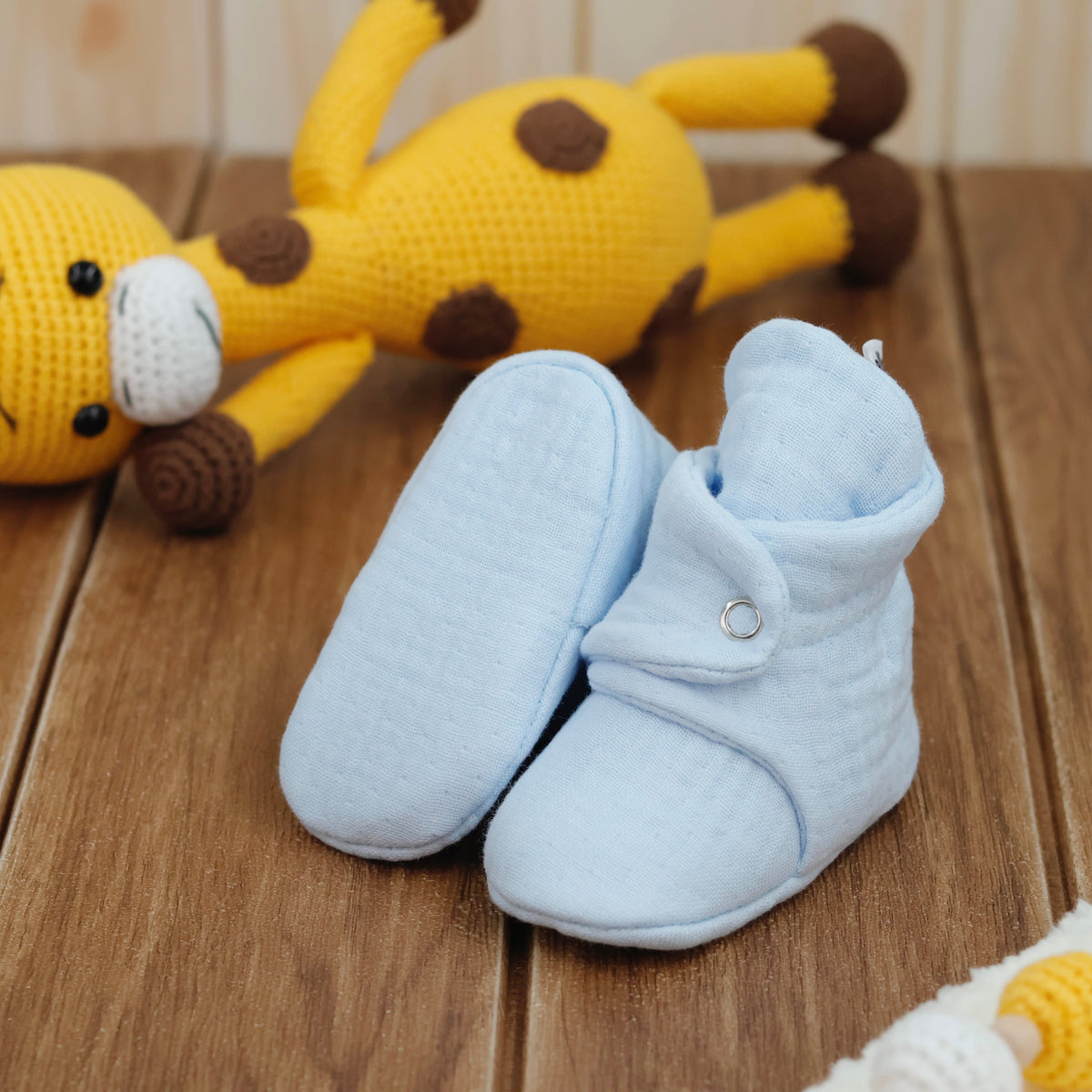Light blue textured muslin baby booties with snap straps arranged on a surface with one sole facing the camera.