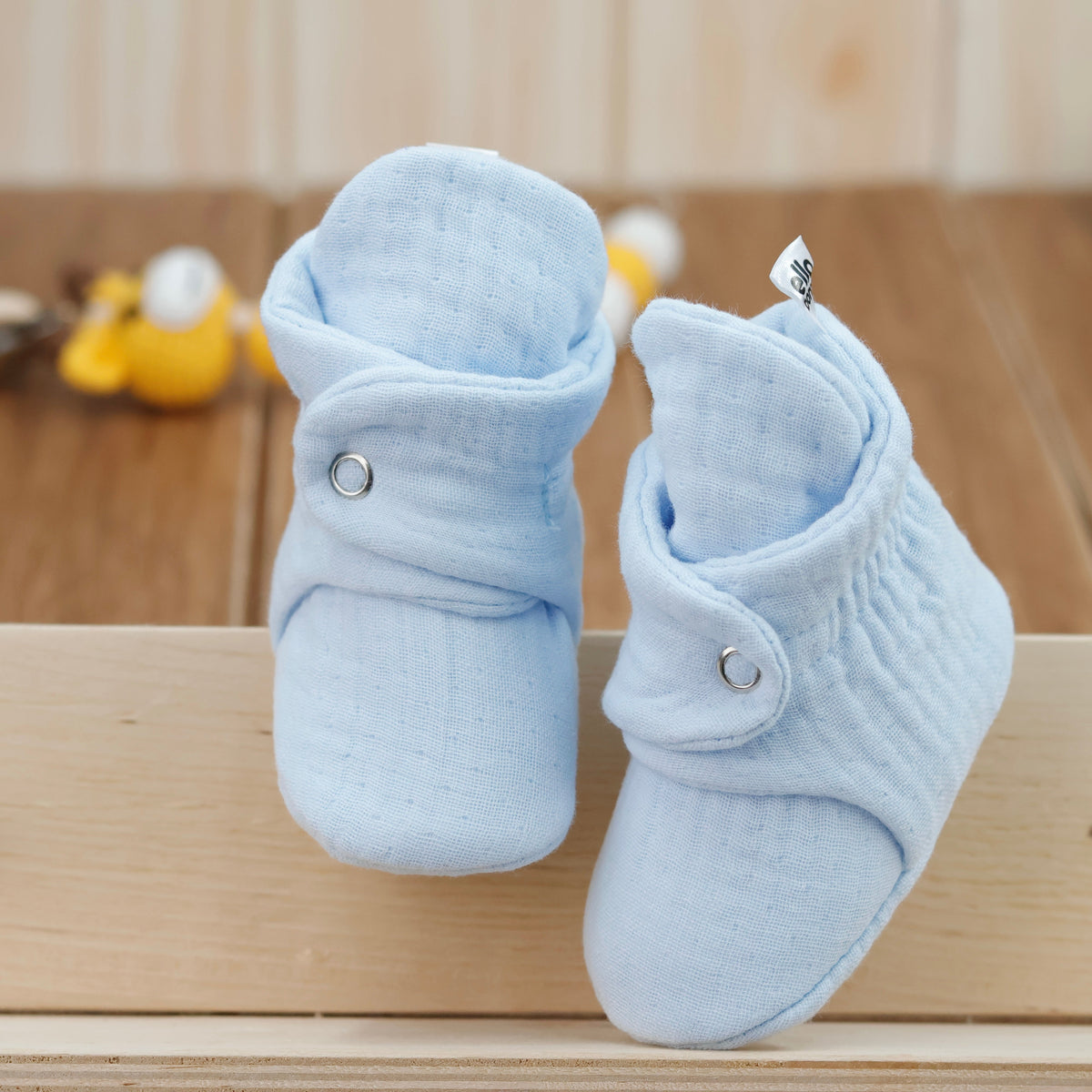 Front view of a pair of light blue textured muslin baby booties with snap straps.