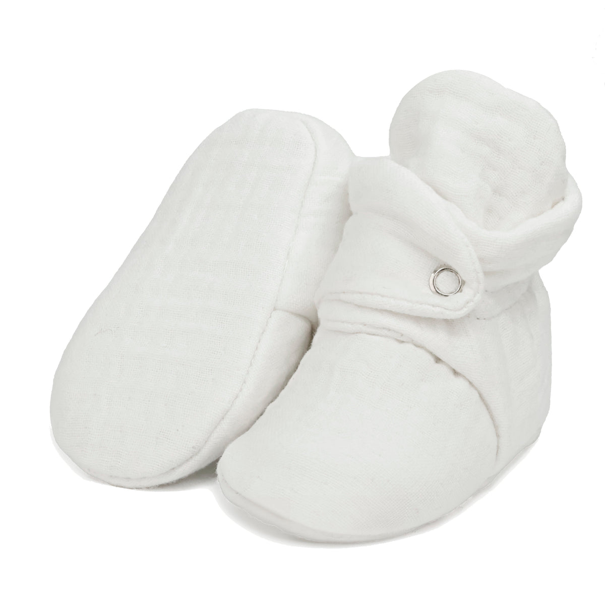 Cream textured muslin baby booties with snap straps, with one sole facing the camera on a white background.