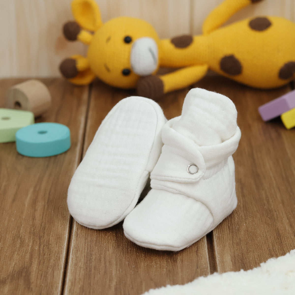 Cream textured muslin baby booties with snap straps arranged on a surface with one sole facing the camera.