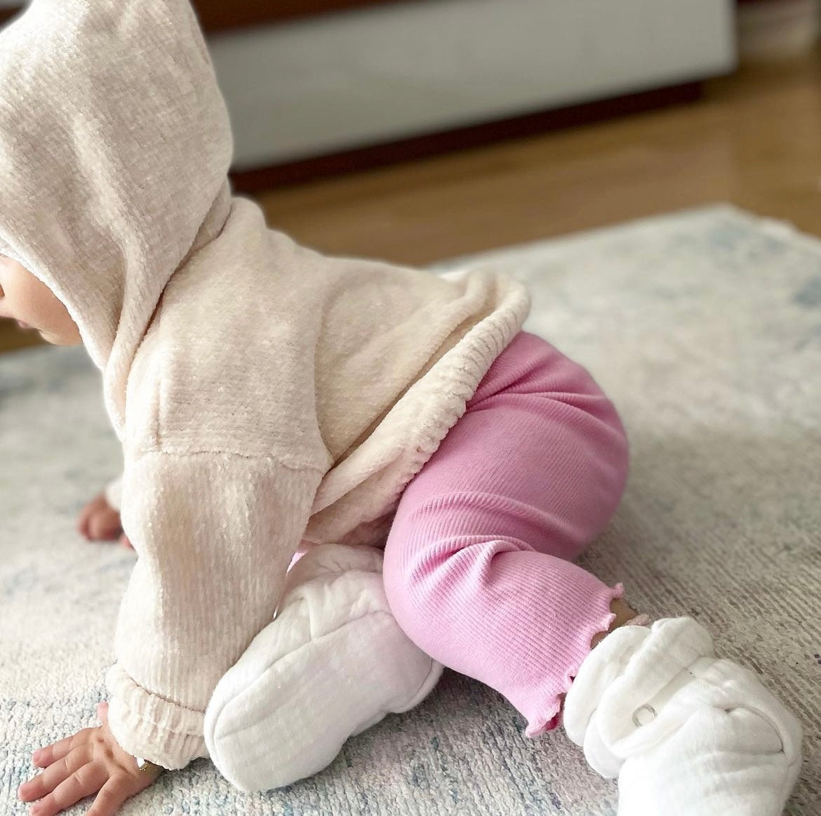 Baby seated on a rug wearing cream booties and pink trousers, side view.