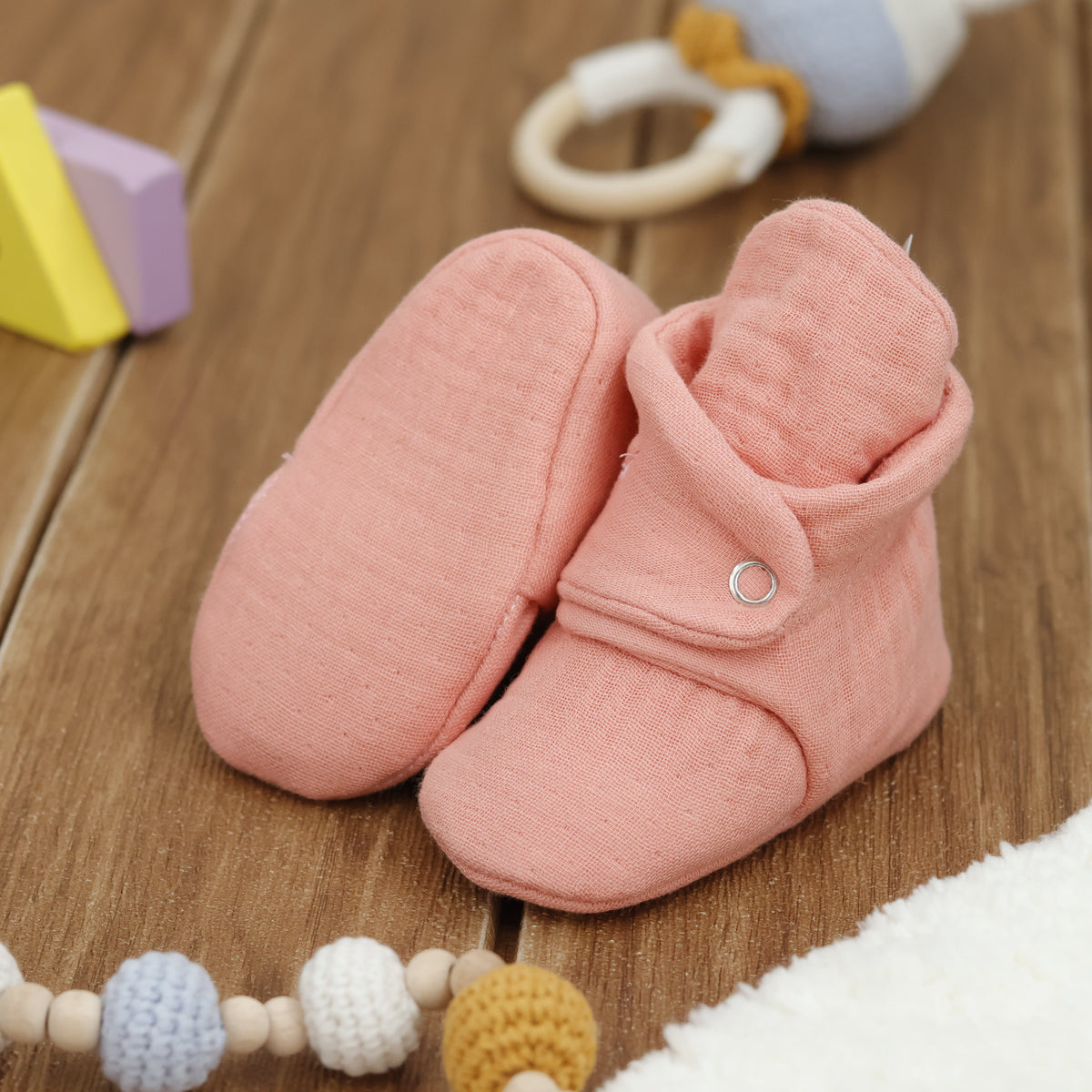 Dusty coral pink textured muslin baby booties with snap straps arranged on a surface with one sole facing the camera.